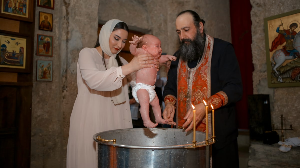 A baby being baptized by a priest and woman in a traditional church setting.