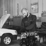 Monochrome photo of a mechanic inspecting a car engine inside a garage. Tools visible in background.
