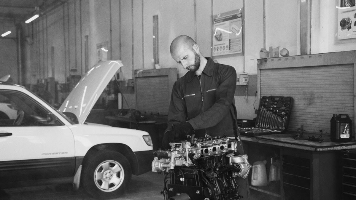 Monochrome photo of a mechanic inspecting a car engine inside a garage. Tools visible in background.